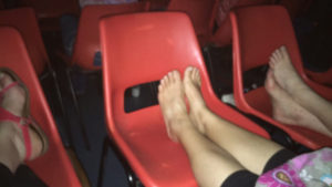 photograph of 3 sets of feet, bare and in sandals, propped up on plastic orange chairs