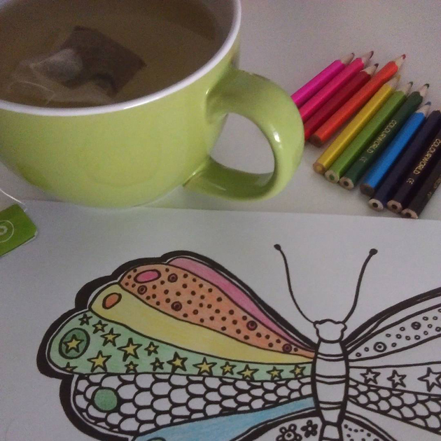 cup of tea and coloured pencils alongside line drawing of butterfly being coloured in rainbow-like