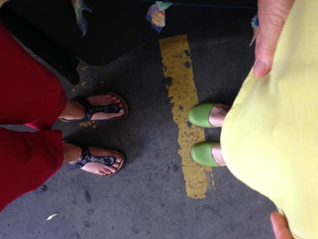2 people looking down at their feet in a carpark; the person on the left has red pants and black sandals with flowers, the person on the right has yellow clothing and green shoes. On the ground is a worn yellow painted car park divider line.