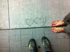 pair of feet in walking boots, pair of feet in sandals, standing on bricks and facing 2 hearts found in concrete