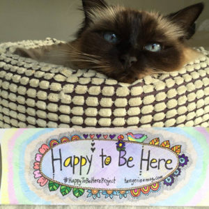 birman ragoll cat resting it's head on a basket, with Happy To Be Here project bookmark in foreground