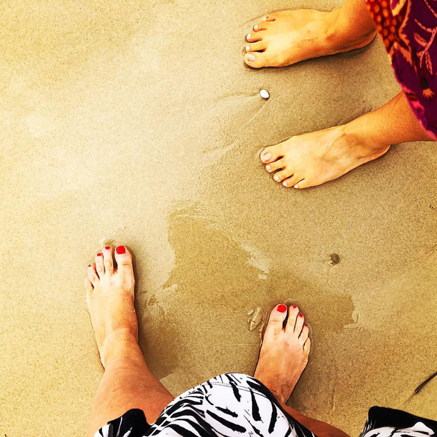 bare feet of 2 humans in skirts, the one taking the photo has applied nail polish, the feet are at a sandy beach