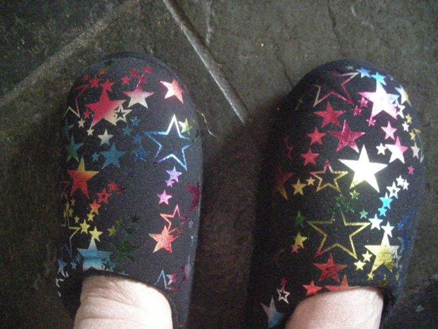 feet with slippers on dark grey slate floor. The slippers have a shiny star pattern in rainbow colours
