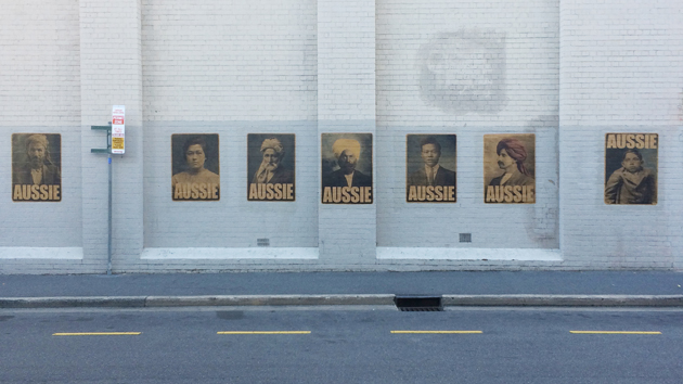 full set of 7 AUSSIE posters by Peter Drew Arts, pasted on a white wall with the road showing in the foreground, with yellow dashed painted markings