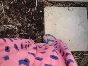Person wearing a pink spotty dress and blue sandals standing on a paving stove near some brown earth