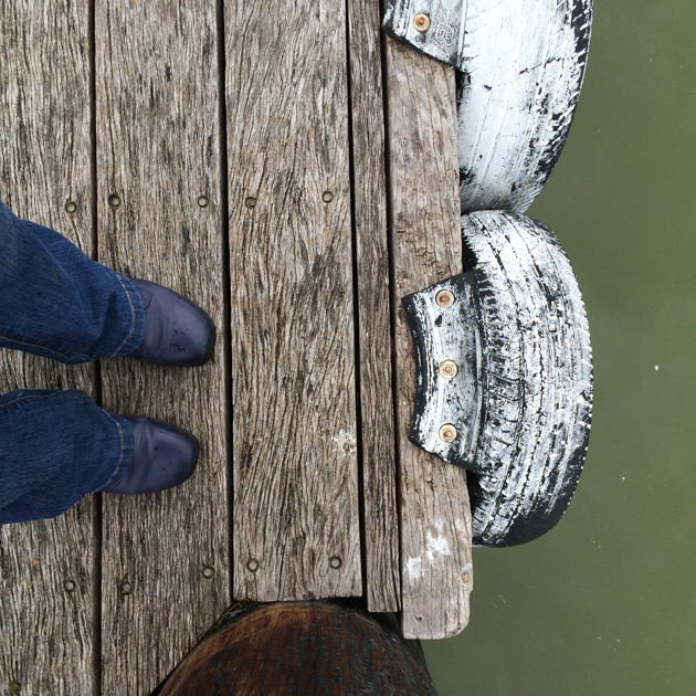 looking downwards at blue jeans and shoes standing on wooden wharf with white painted tyres attached to the edge and green water below