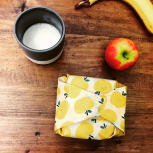 A cup of milk, an apple, a banana and a snack wrapped in a yellow apple patterned food wrap, on a wooden surface.