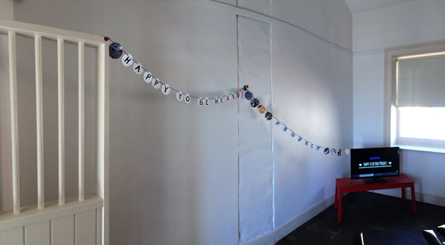white room with circle bunting leading from a white barred door to a TV on a red bench. Both the bunting and the screen shot spell out: Happy To Be Here Project.