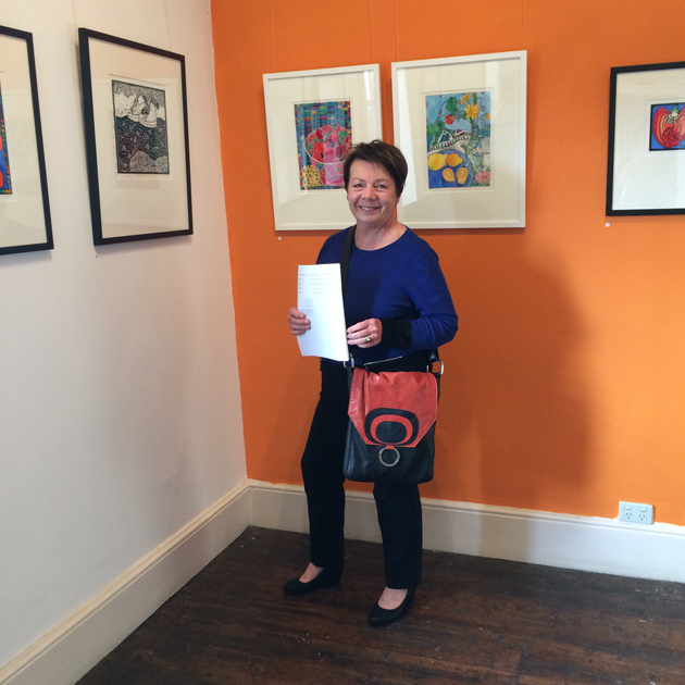 Smiling woman in blue top and holding red and black graphic design bag, standing near art exhibition works hung on 2 walls, one wall of which is painted orange.