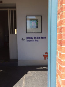 Looking into a red brick building through a blue door frame, is seen a white wall with a painting displayed. Underneath the picture are the words "Happy To Be Here" and "Tangerine Meg" with an arrow pointing left, and a sticker of an orange cat perched just above the skirting board.