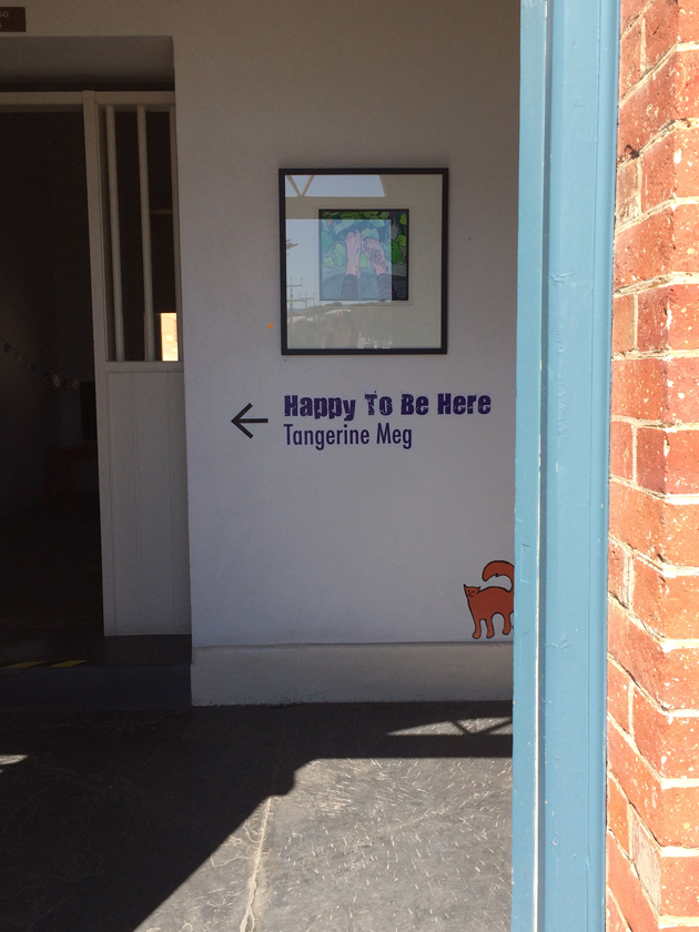 Looking into a red brick building through a blue door frame, is seen a white wall with a painting displayed. Underneath the picture are the words "Happy To Be Here" and "Tangerine Meg" with an arrow pointing left, and a sticker of an orange cat perched just above the skirting board.