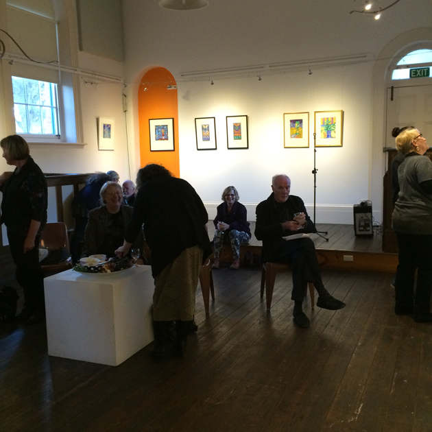delightful bold souls mingling, chatting and smiling at the launch of Happy to Be Here exhibition; the room is high of ceiling, with walls lined with framed pictures; the floor is dark timber.