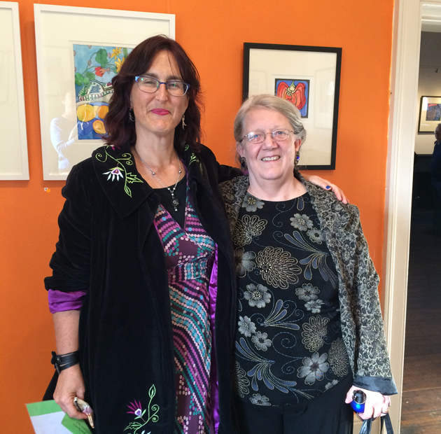 Tangerine Meg and author Susan Fitzgerald standing in fromt of orange wall displaying framed brightly coloured lino prints and watercolours