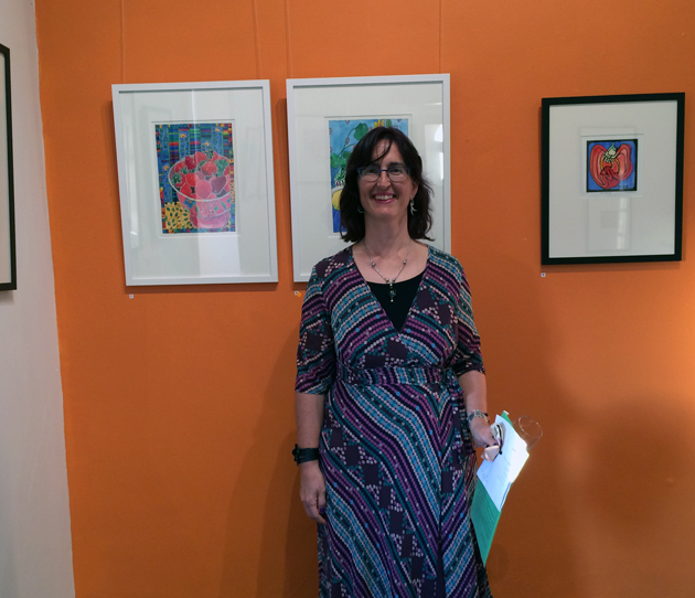 South Australian woman artist, Tangerine Meg, wears a purple and green/blue spotty dress, and stands in front of an orange wall with framed pictures hanging on it.