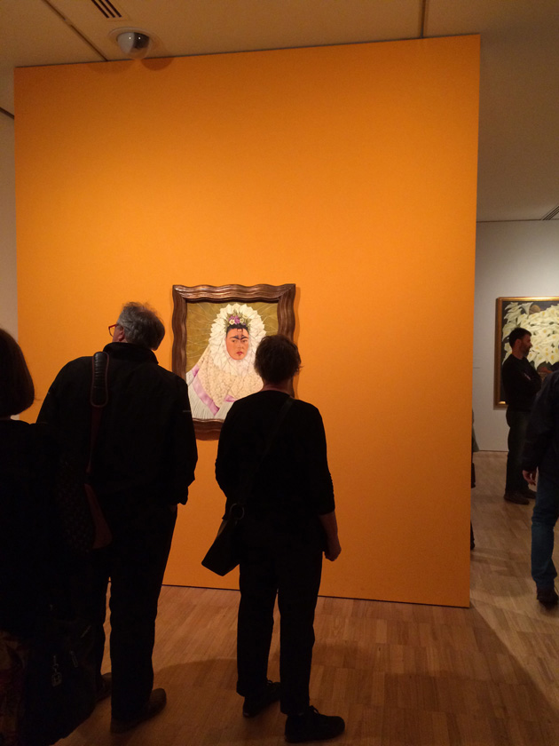 Backs of people viewing Frida Kahlo painting on orange wall.