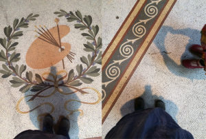 Feet standing on mosaic floor. The floor has small tiles making a curved repeated pattern and featuring an artist's palette surrounded by an olive branch design.