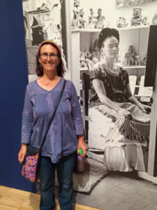 Tangerine Meg posing smiling in blue shirt in front of black and white photo mural of Frida Kahlo in her studio.