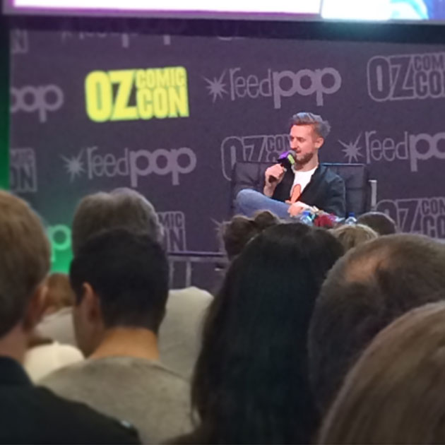 Audience members facing away from the camera, towards a man with a microphone talking on stage. The man's backdrop is patterned with "Oz Comic Con" and "reed pop" logos.