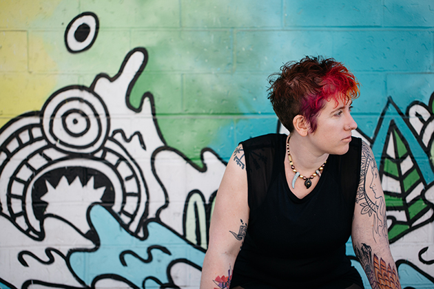 Michelle Nickolaisen seated in front of a graffiti wall with black drawings and lots of green and blue. She's looking out of the photo to our right.